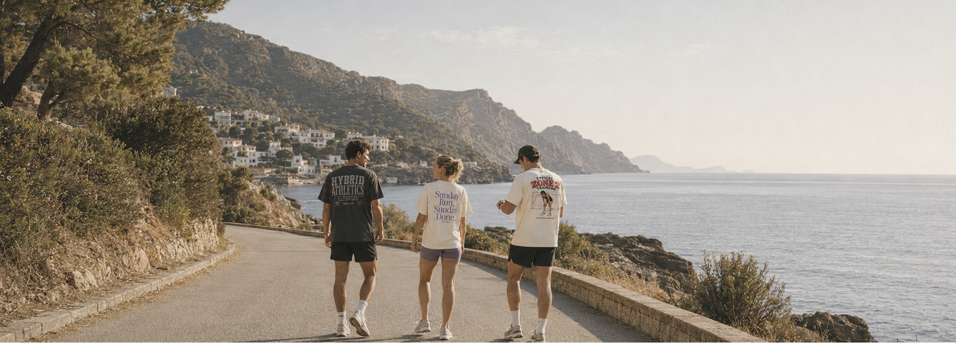 Three people walking along a scenic coastal road with mountains and sea in the background.